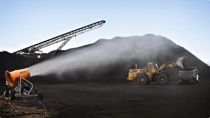 Dust cannon throwing atomized mist at coal stockpiles with machinery unloading