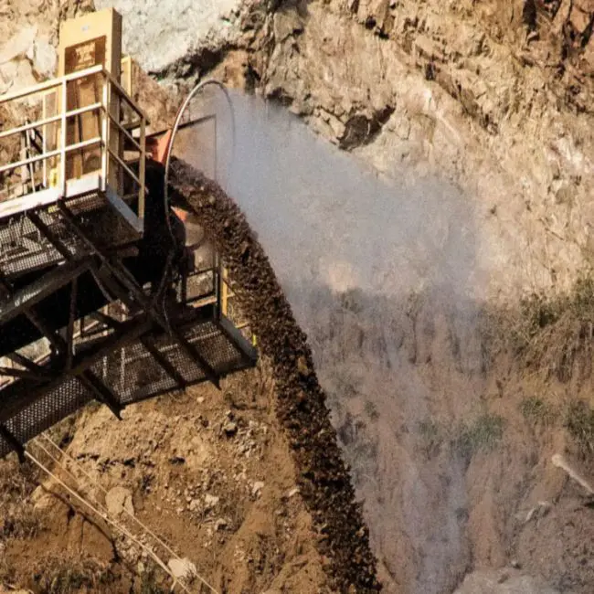 A large conveyor belt pours rocks down a dusty quarry slope with a water spray in action in the USA.