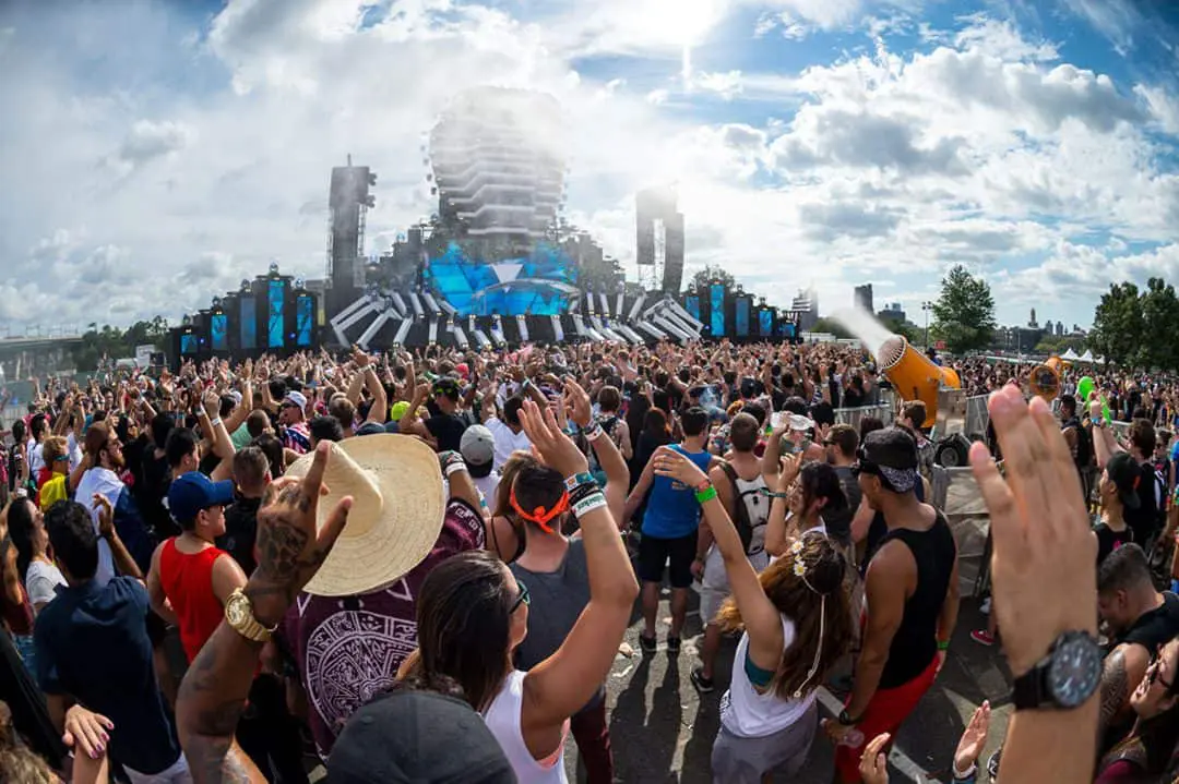 A large crowd of diverse people is enjoying a vibrant music festival under a clear blue sky